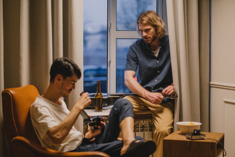 Two men relaxing at home, enjoying drinks and a guitar by the window.