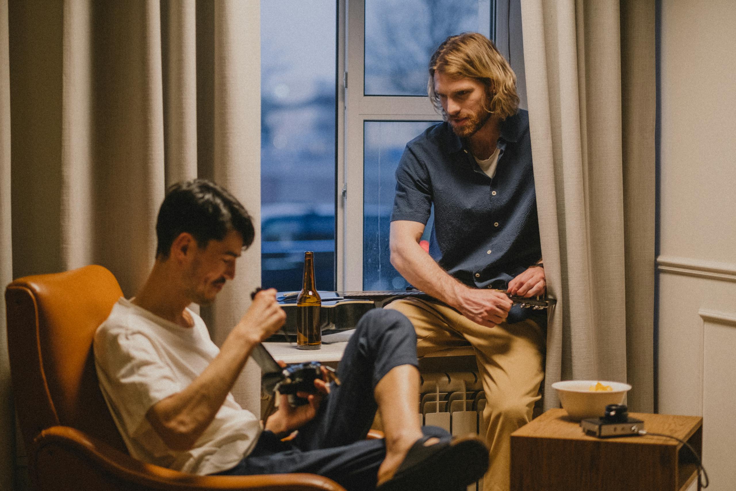 Two men relaxing at home, enjoying drinks and a guitar by the window.