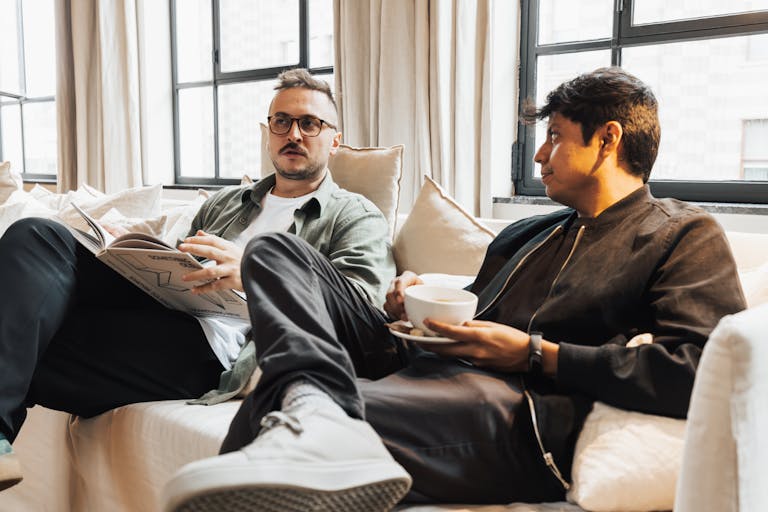 Two men sitting on a cozy sofa, one reading a magazine, enjoying coffee in a bright living room.