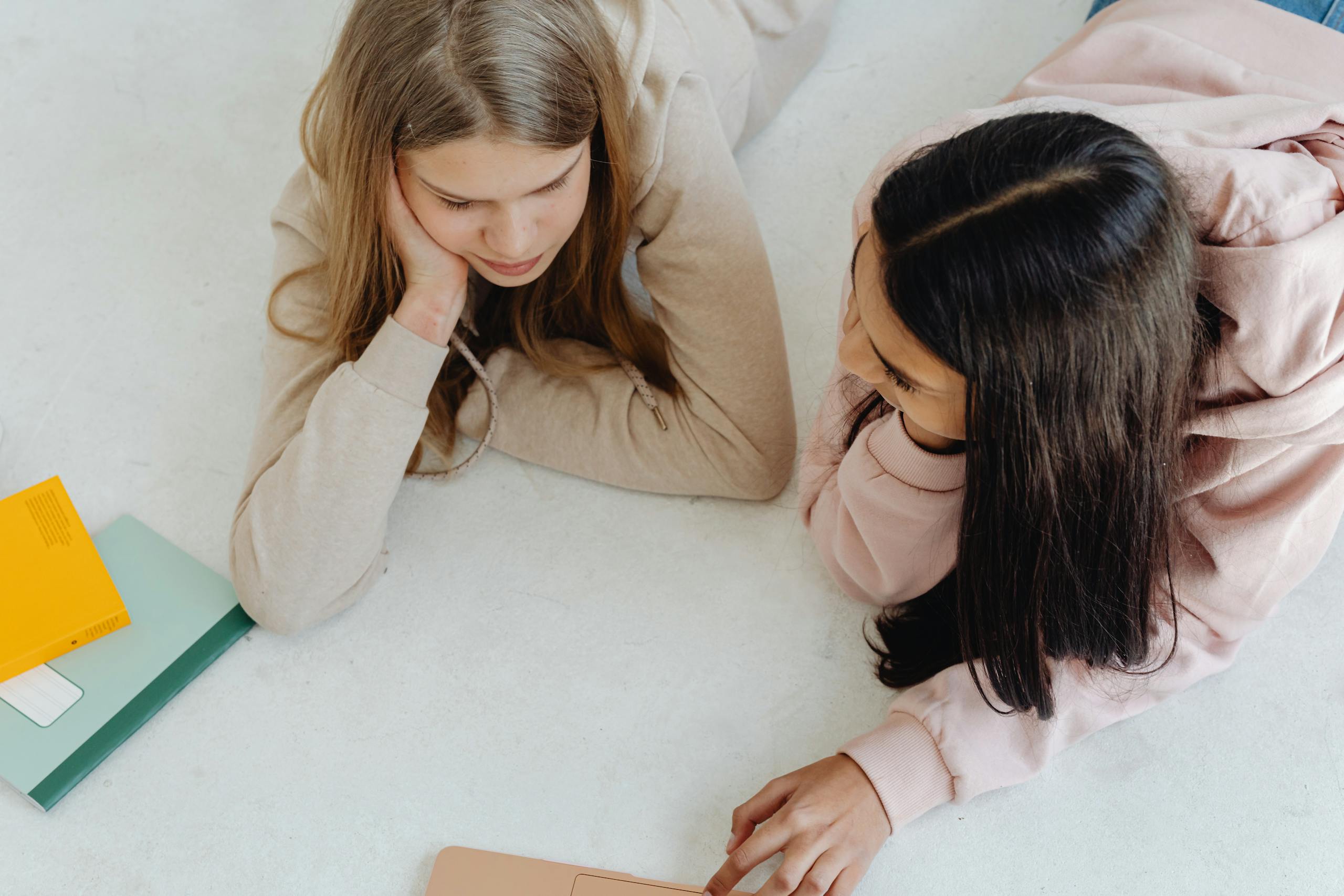 Two teenage girls lying on the floor looking at a laptop in a cozy indoor setting.