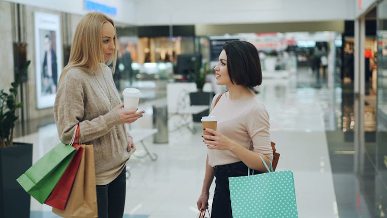 Two women enjoy coffee while shopping inside a stylish mall.