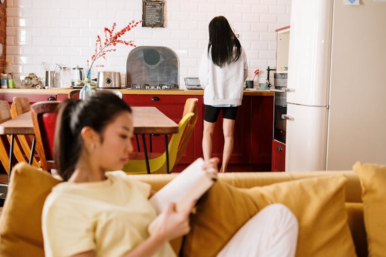 Two women relax at home. One reads on the sofa while the other is at the kitchen counter.