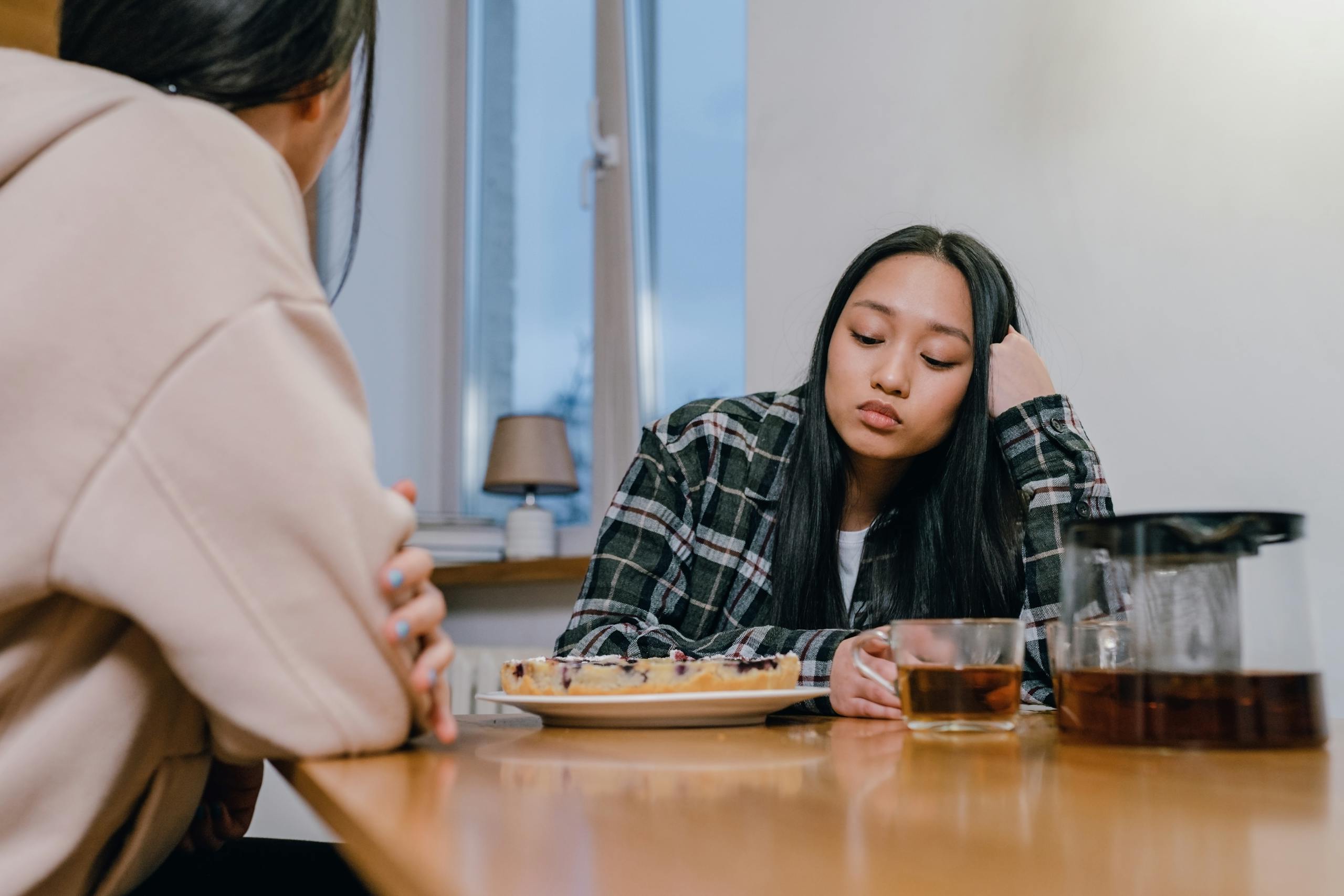 Two women sit at a table indoors, quietly sharing a moment, one looks thoughtful.