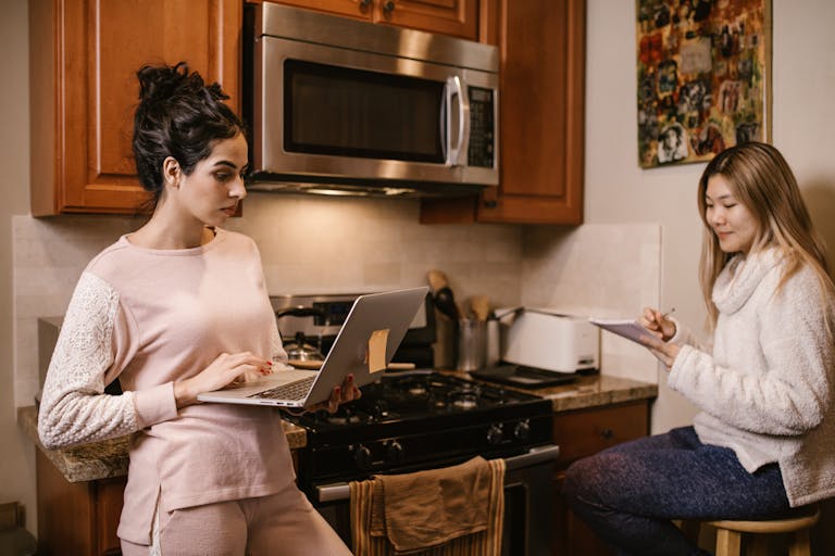Two women working from home in the kitchen with a laptop and notebook, focused and serious.