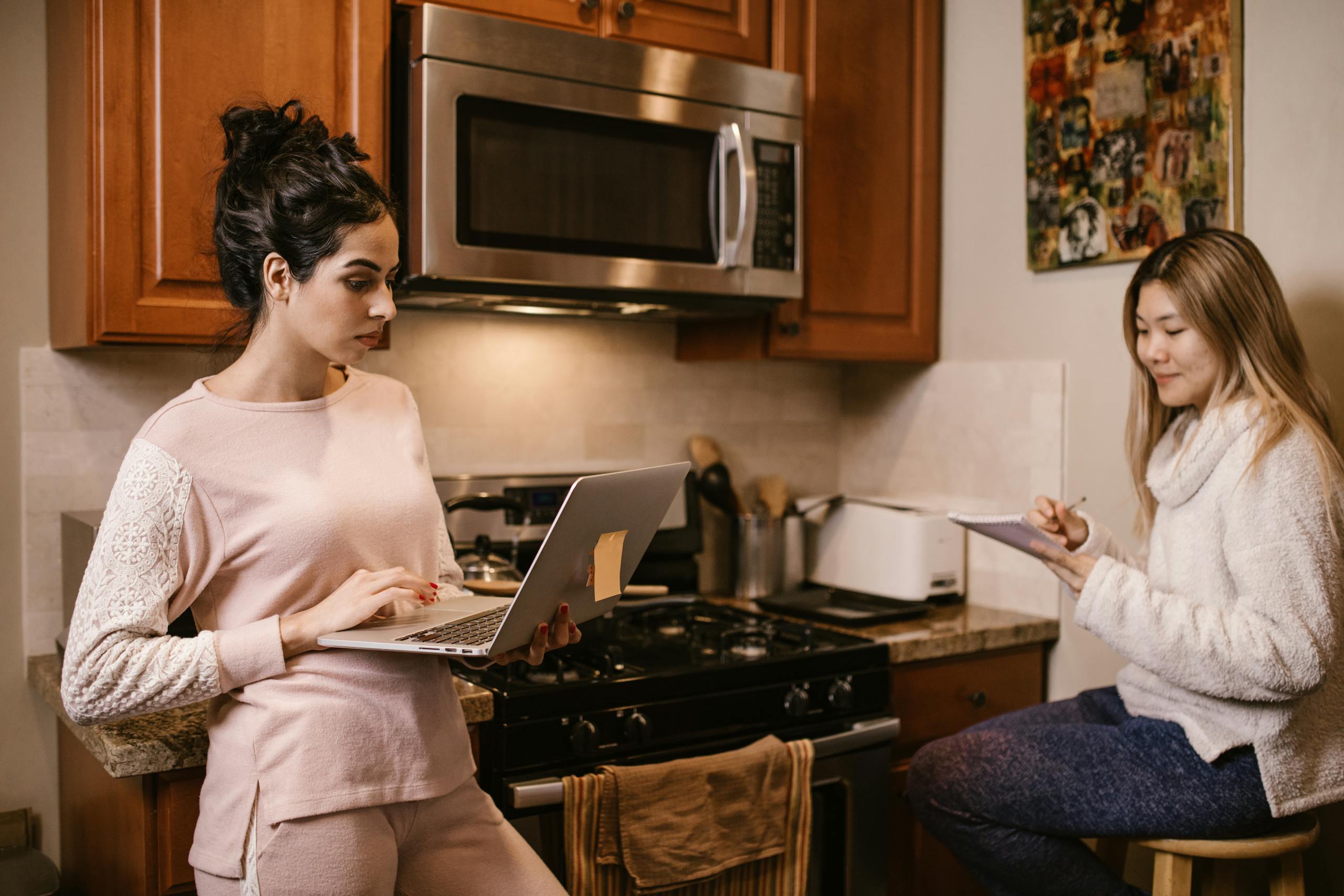 Two women working from home in the kitchen with a laptop and notebook, focused and serious.