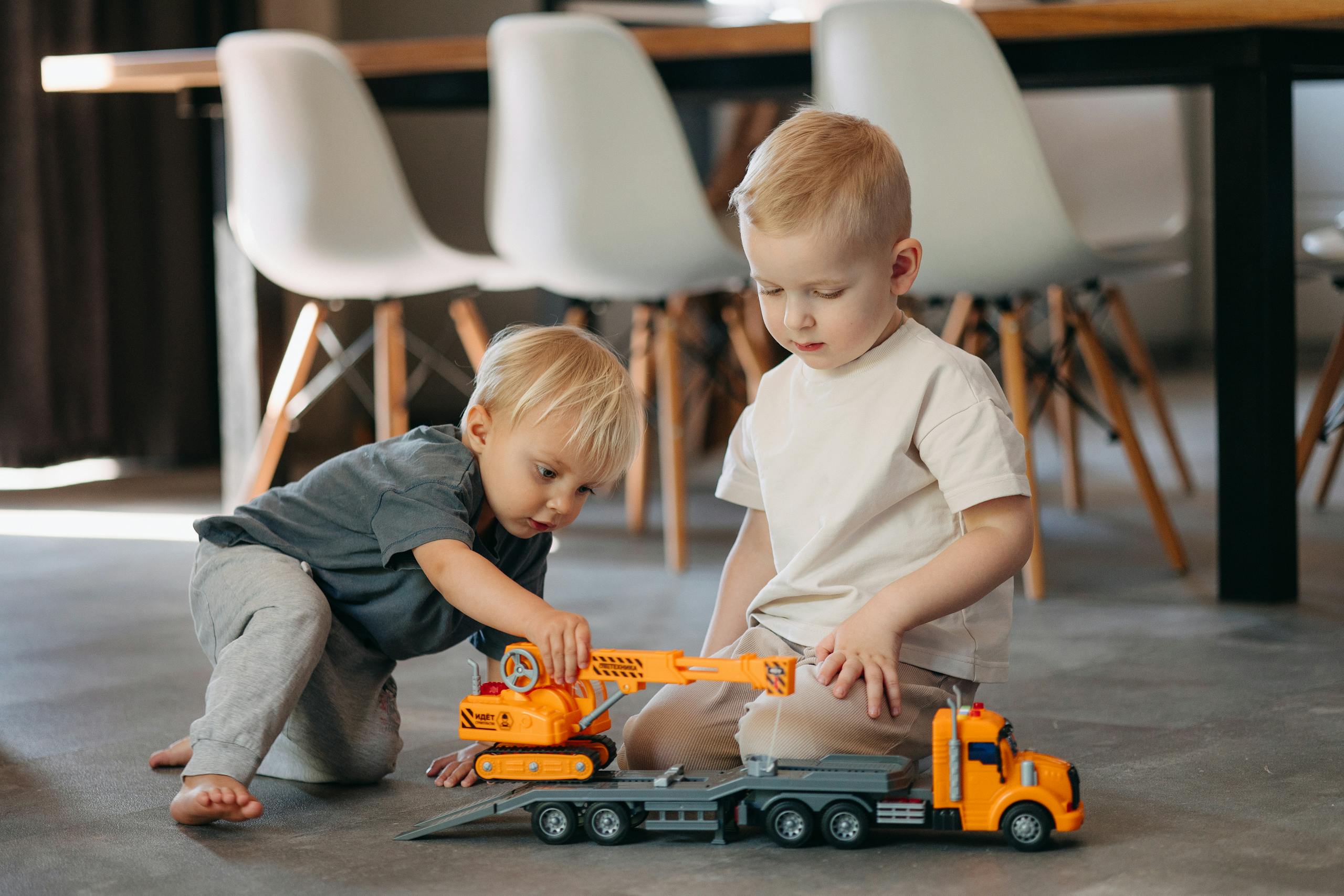 Two young boys playing with toy trucks on the floor in a modern indoor setting.