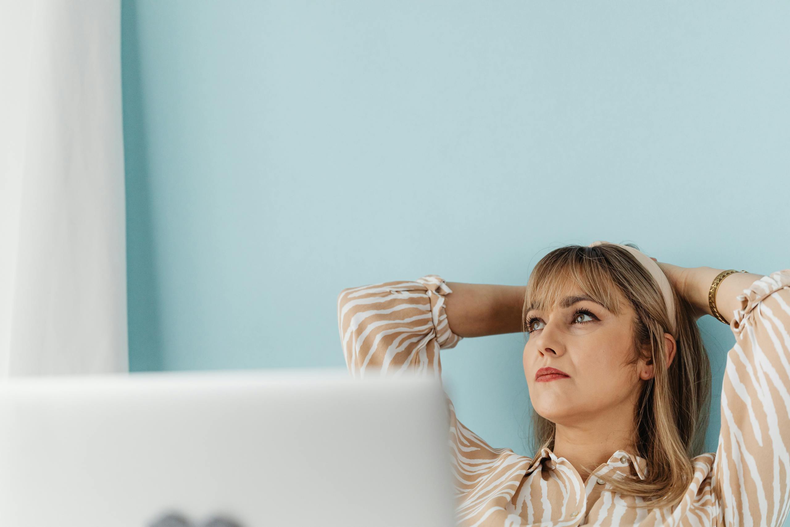 Woman in a striped blouse sitting with hands behind her head, contemplating while working remotely.