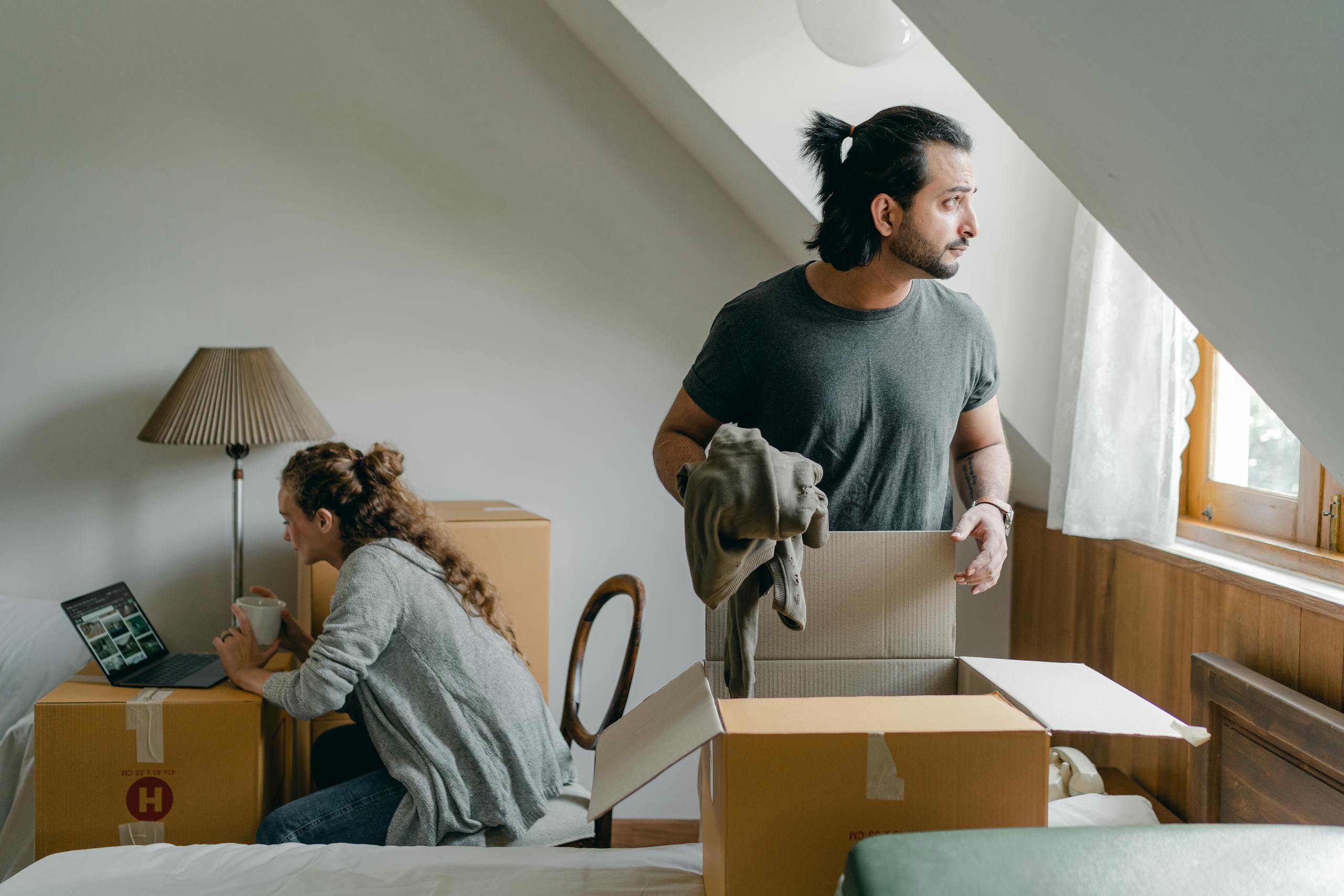 Woman in casual wear drinking coffee while using social media on laptop and sitting on chair near thoughtful ethnic partner getting sweater out of cardboard box in new house
