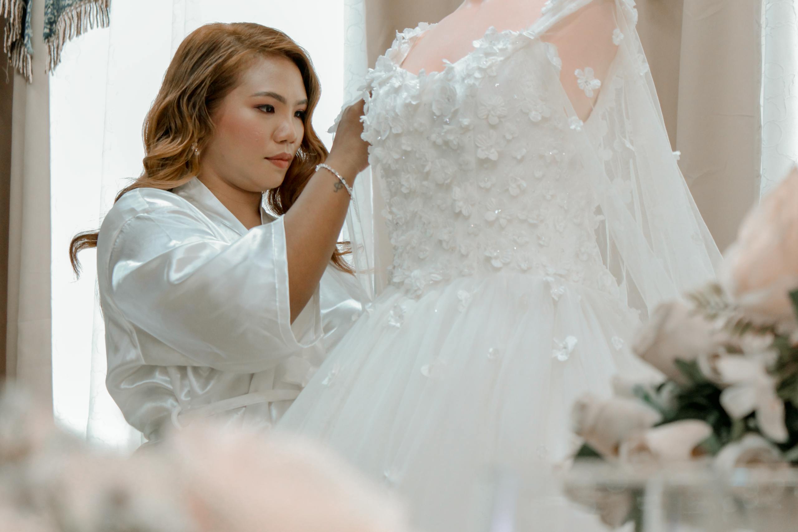 Woman in white robe carefully adjusts detailed wedding dress indoors.