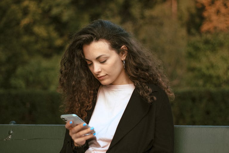 Woman with curly hair checking smartphone while sitting outdoors in Warsaw park.
