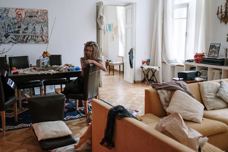 Woman with dreadlocks sitting in a cozy, cluttered room checking her smartphone.