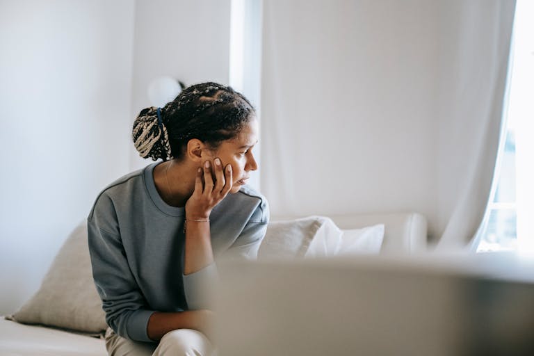 Young black female in casual outfit sitting on cozy white bed and thinking while looking away in daytime