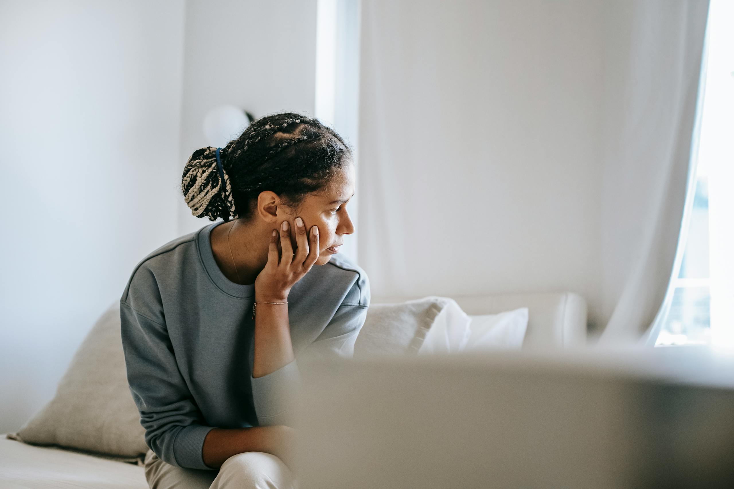 Young black female in casual outfit sitting on cozy white bed and thinking while looking away in daytime