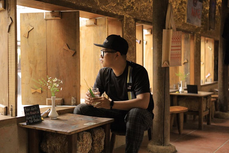 Young man in casual wear sits in a rustic wooden cafe, looking out window thoughtfully.