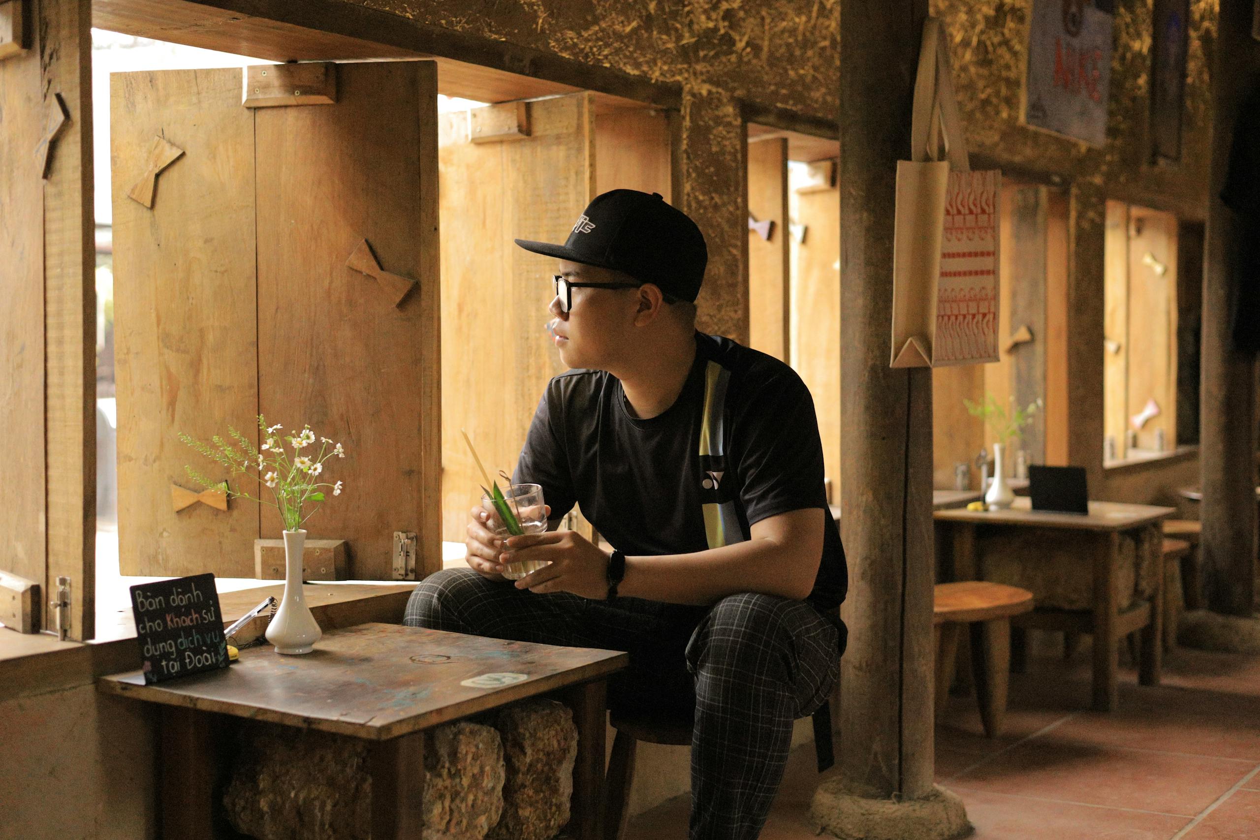 Young man in casual wear sits in a rustic wooden cafe, looking out window thoughtfully.