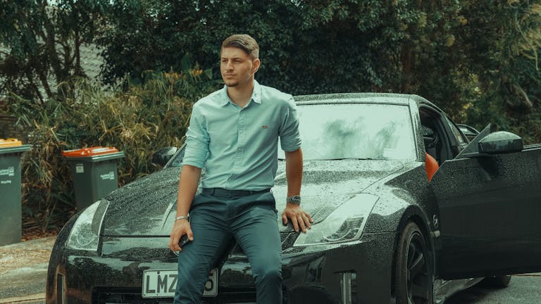 Young man in formal attire leaning on a luxury sports car parked outdoors, exuding style and confidence.