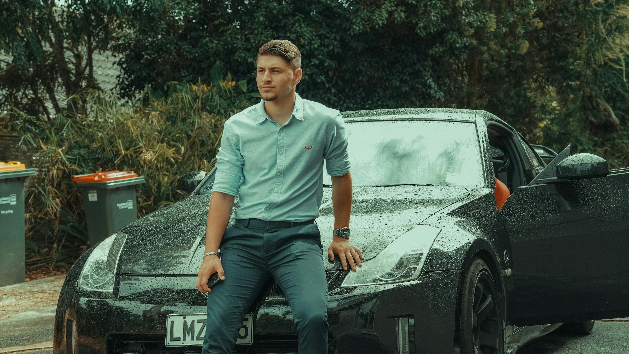 Young man in formal attire leaning on a luxury sports car parked outdoors, exuding style and confidence.