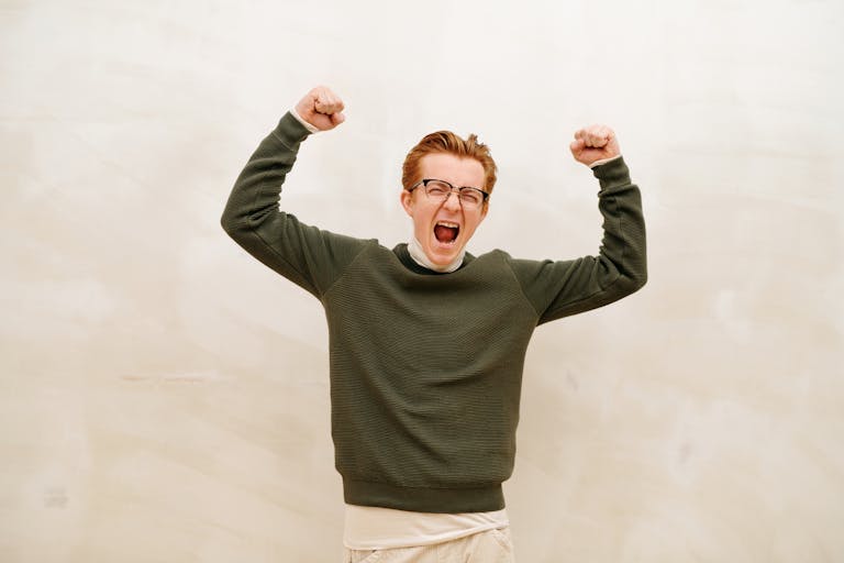 Young man in glasses and sweater jubilantly raises fists indoors, expressing joy and success.