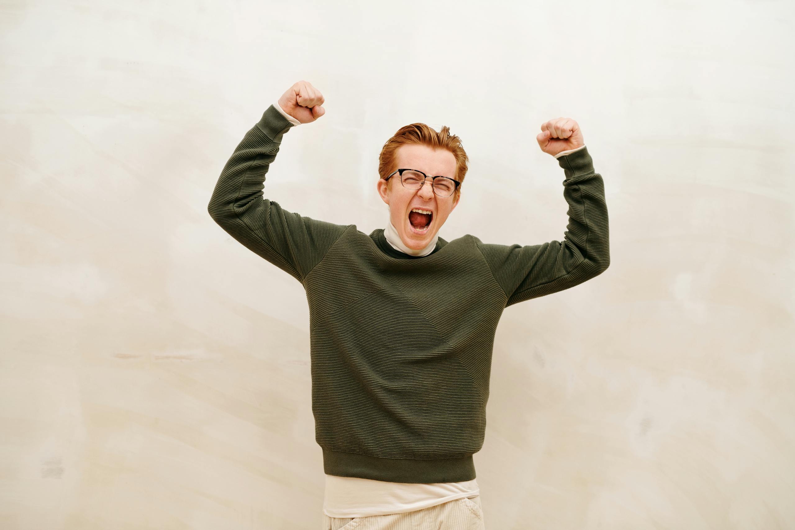 Young man in glasses and sweater jubilantly raises fists indoors, expressing joy and success.