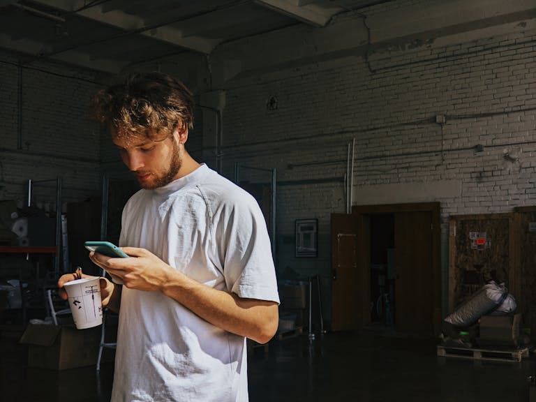 Young man standing in warehouse using smartphone with coffee cup in hand.