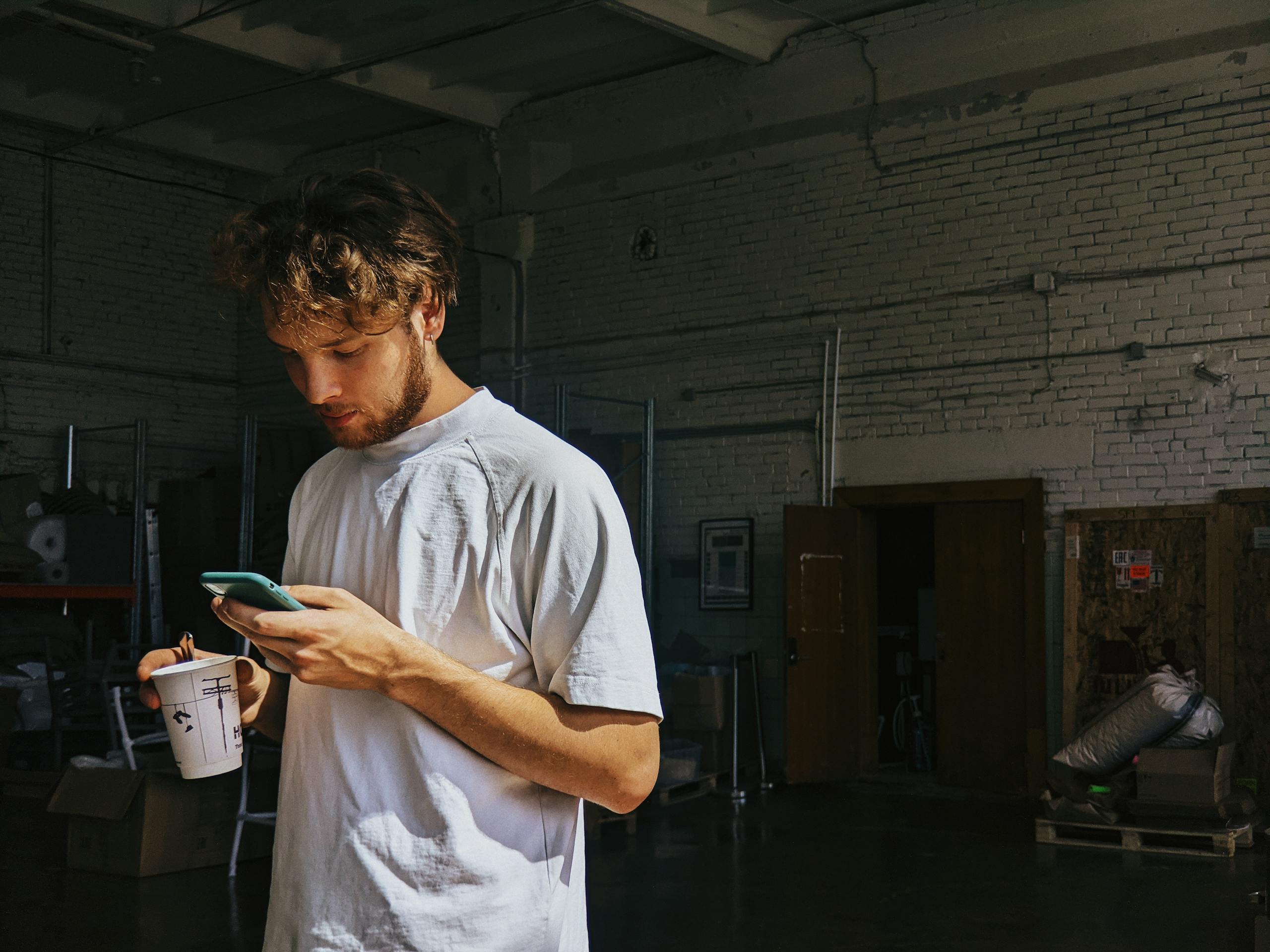 Young man standing in warehouse using smartphone with coffee cup in hand.