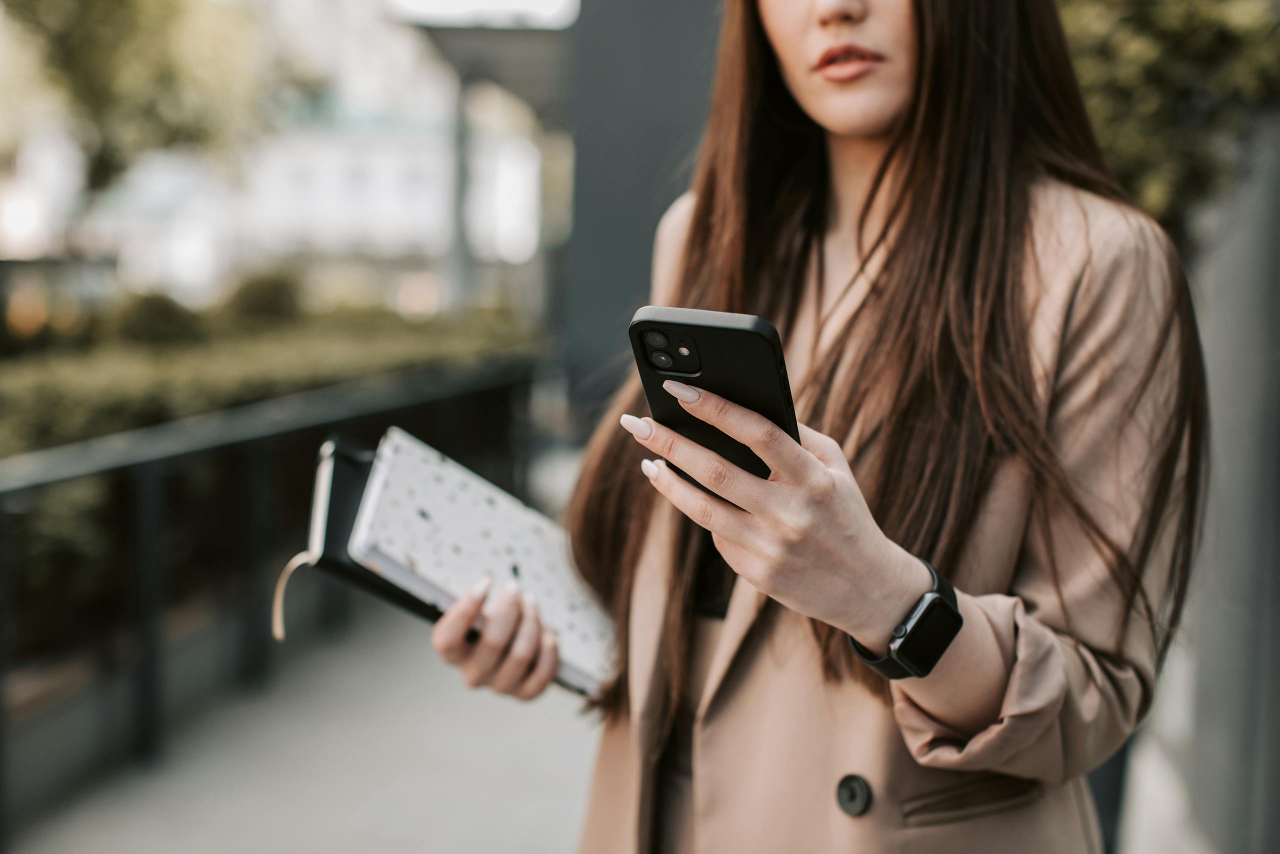 Young woman in beige blazer checking smartphone while holding a notebook outside.