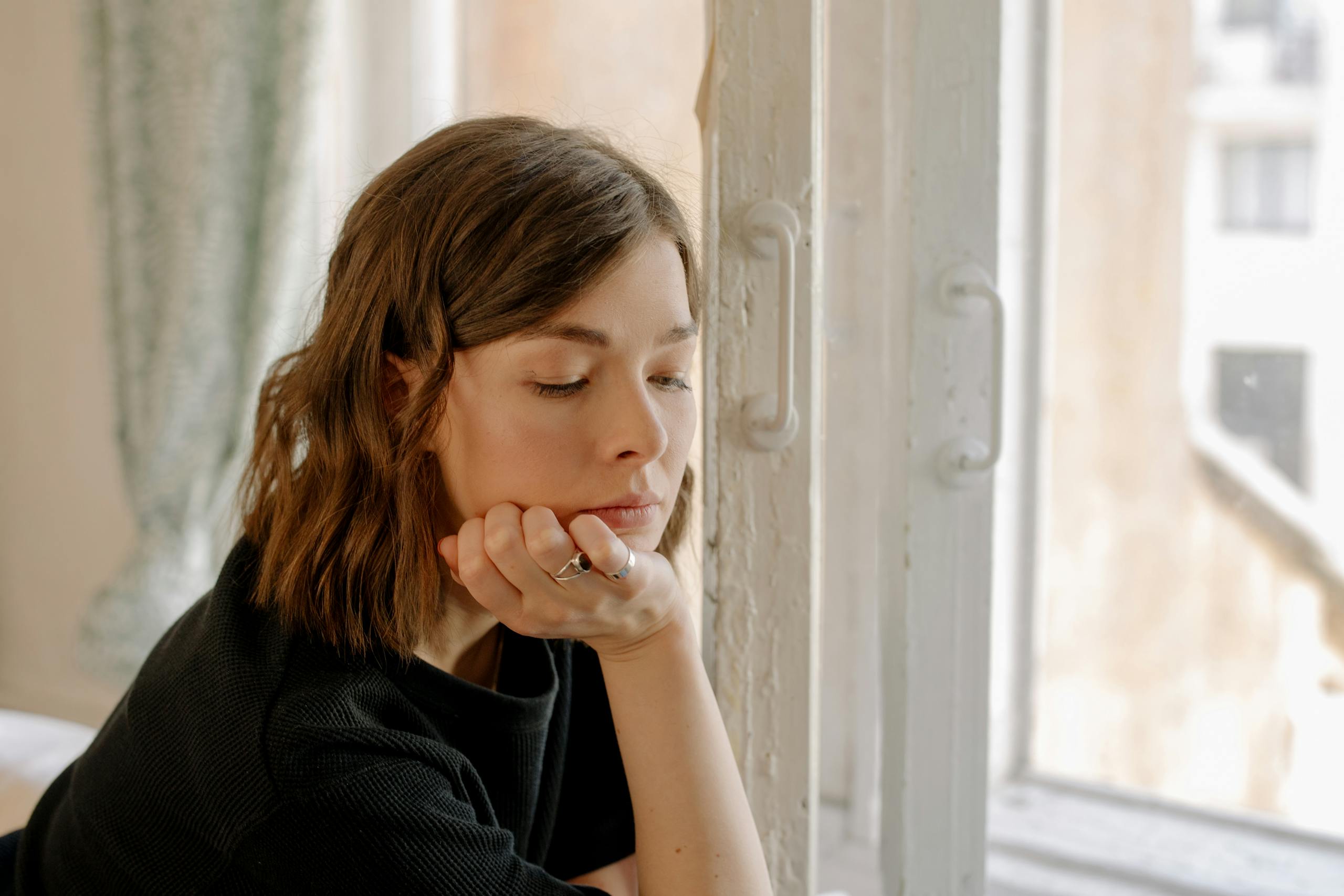 Young woman looking thoughtful while sitting by a window indoors. Natural lighting and contemplative mood.