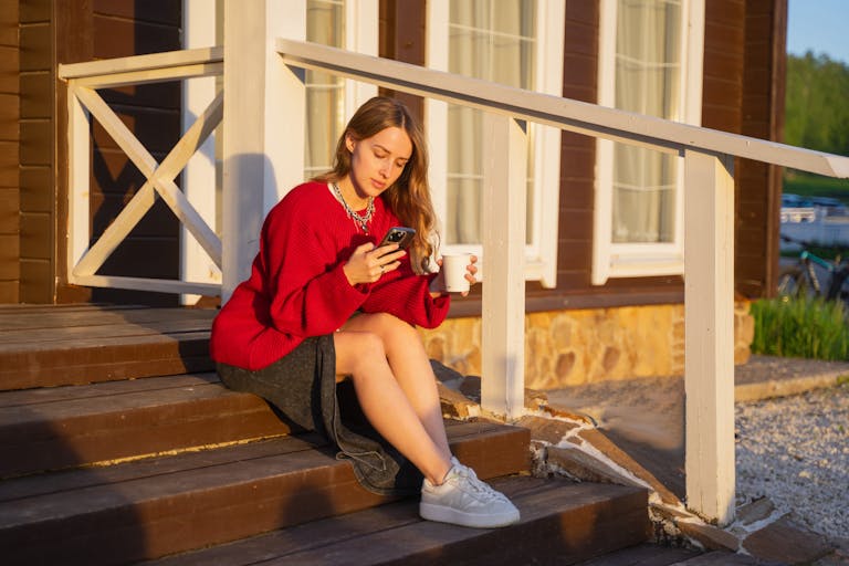 Young woman with a smartphone and coffee on wooden steps, enjoying the sunset.