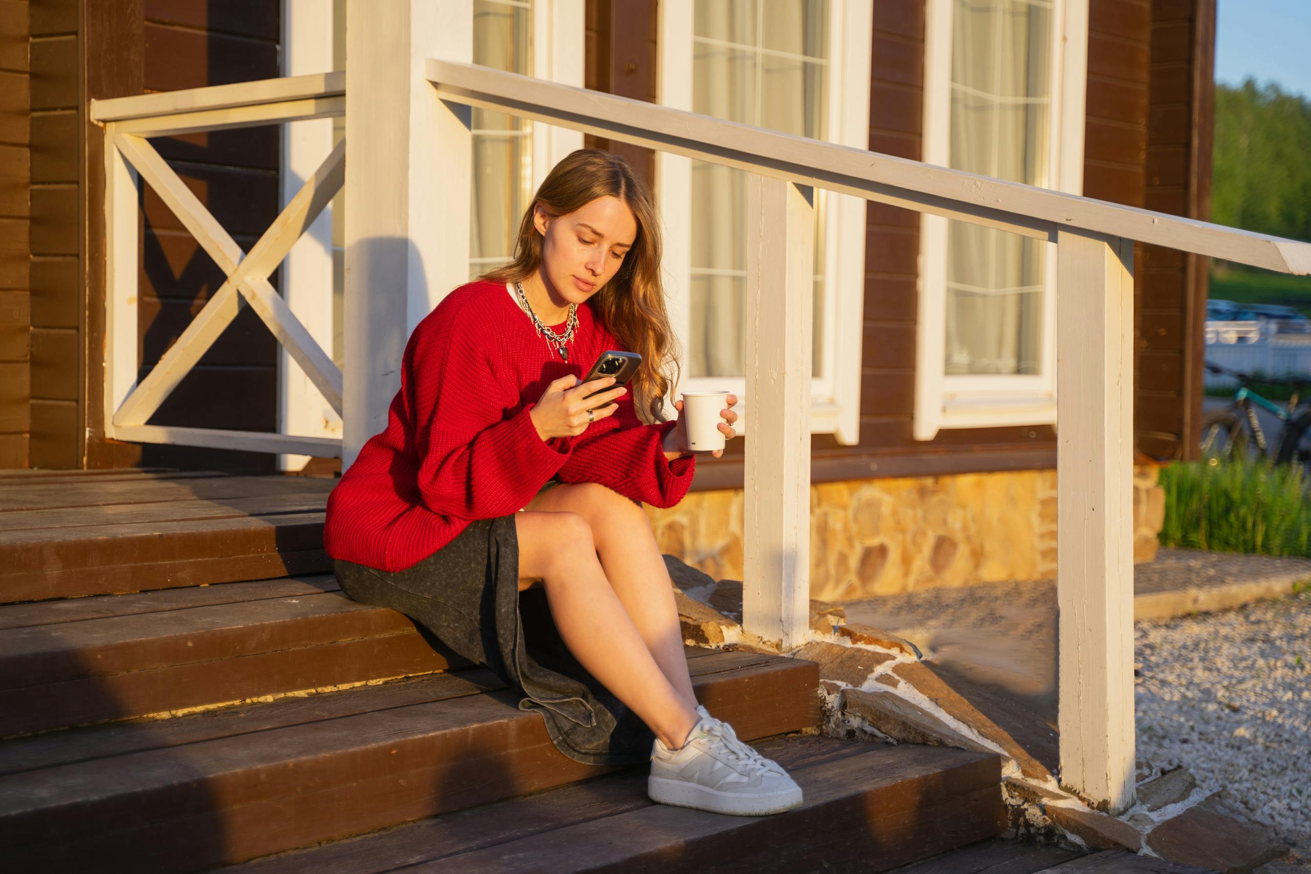 Young woman with a smartphone and coffee on wooden steps, enjoying the sunset.