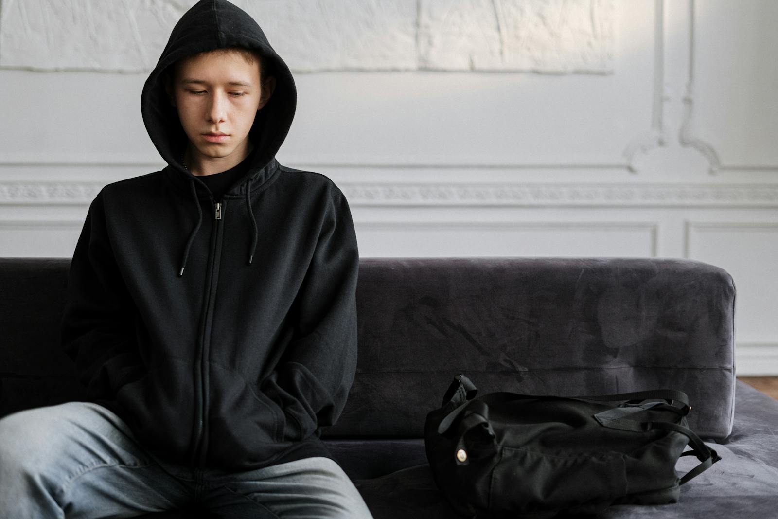 Young teen wearing hoodie sitting pensively on couch indoors.