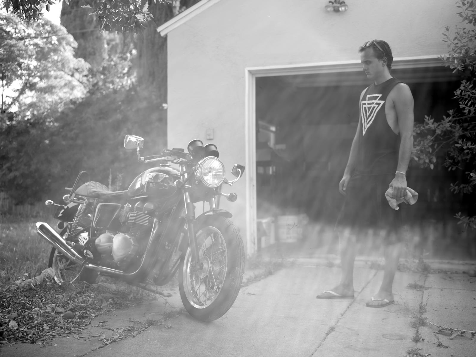 A man stands by a vintage motorcycle in front of a sunny garage, holding a cloth.