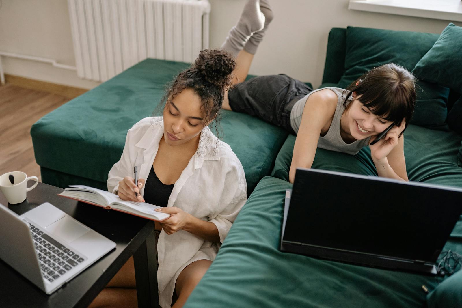 Two women sitting on a green sofa, working on laptops and writing in a notebook, creating a cozy home office vibe.