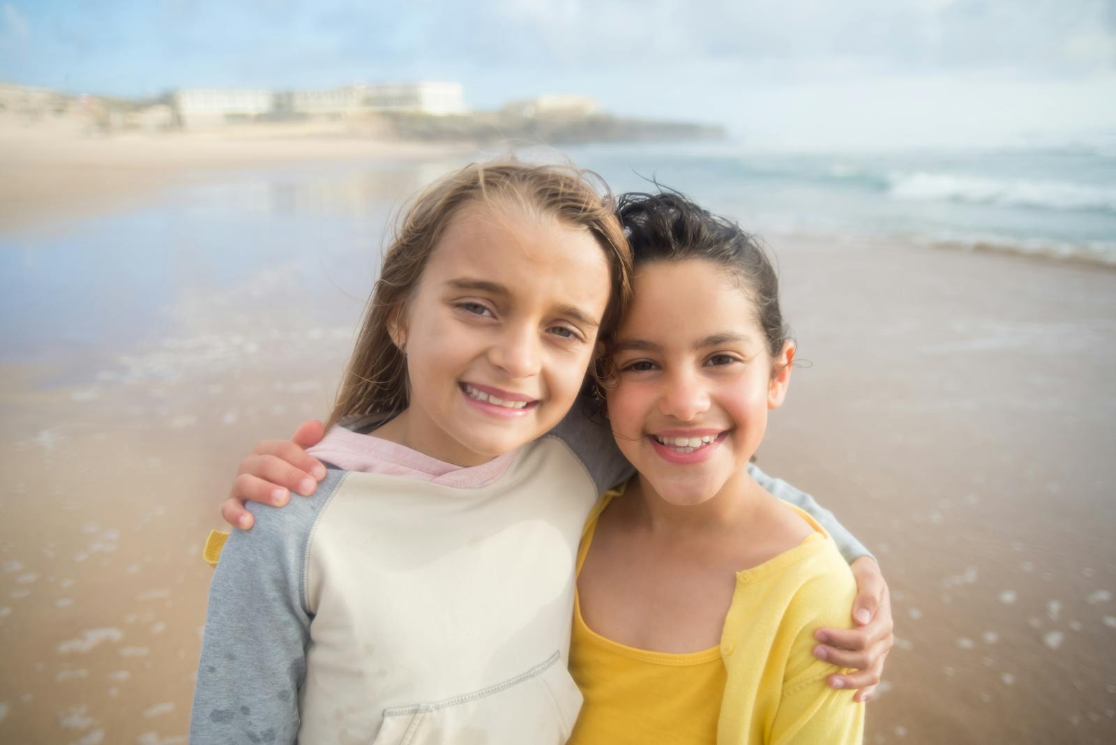 Two happy girls embracing on a sandy beach in Portugal, enjoying a sunny day.