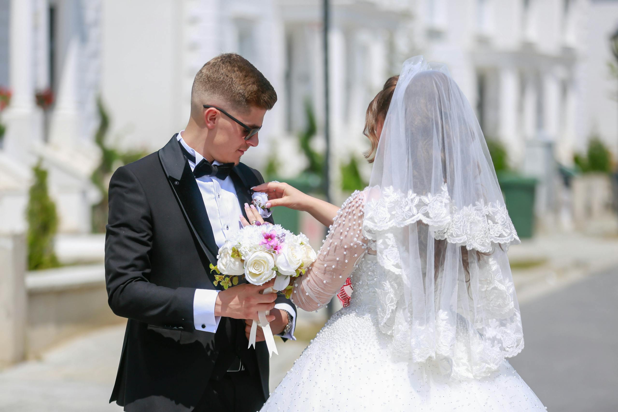 A bride and groom share a moment outdoors on their wedding day, holding a bouquet and dressed in formal attire.