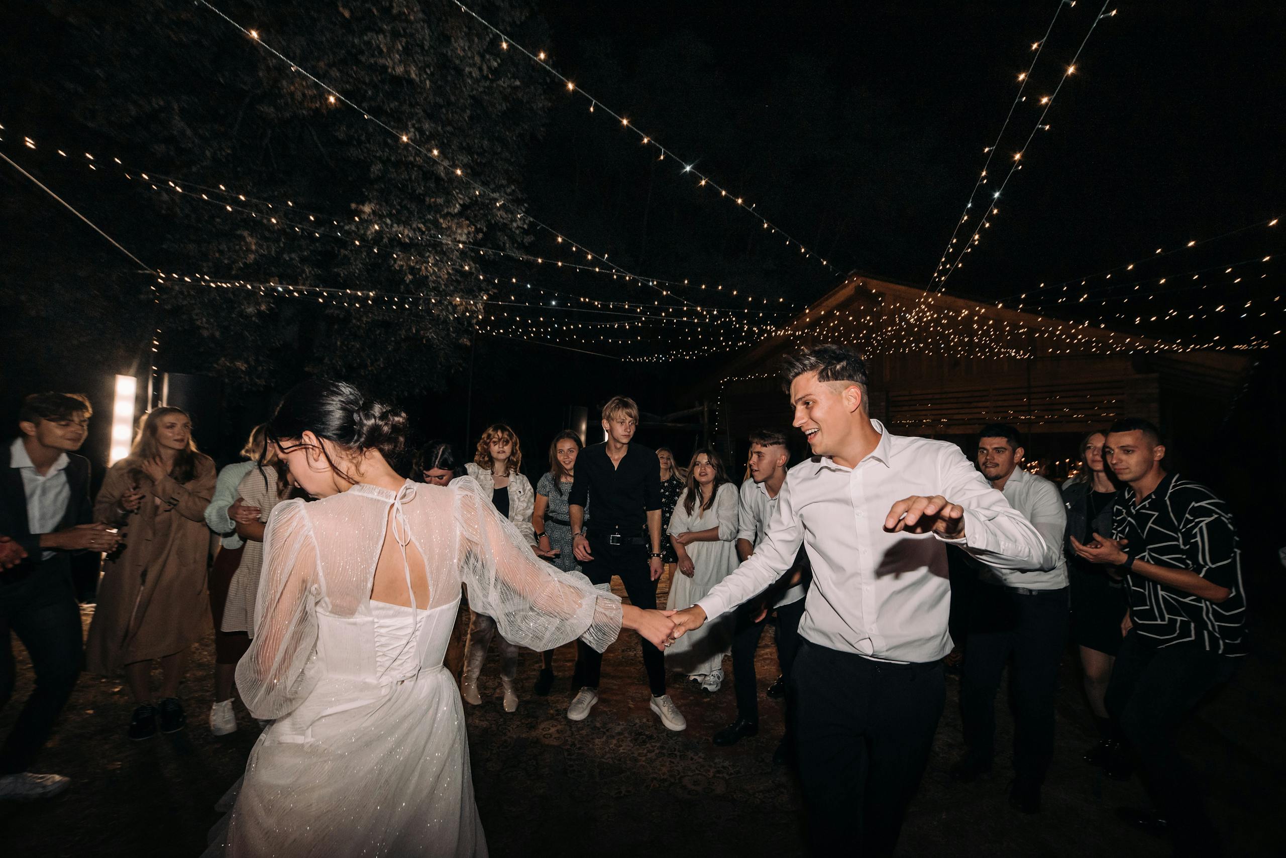 A couple dances joyfully under twinkling lights at an outdoor night wedding celebration.