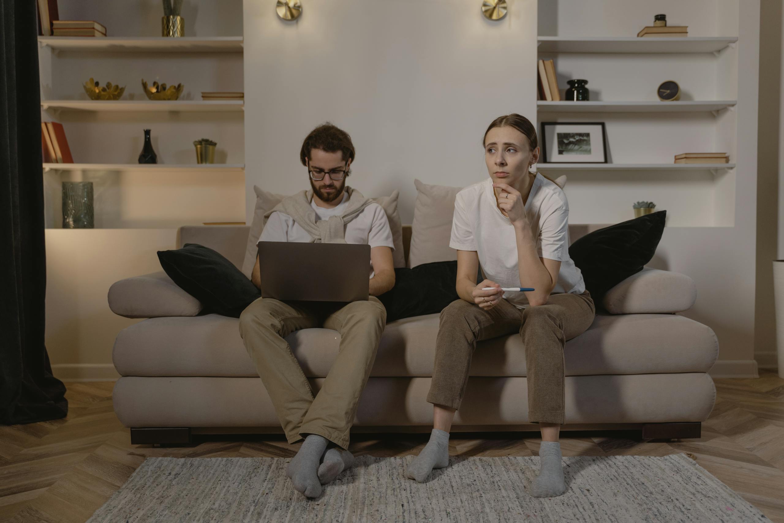 A couple sits on a sofa. The man is using a laptop, while the woman appears thoughtful.