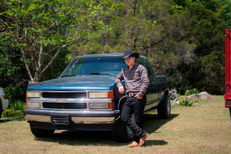 A cowboy in a hat relaxes against a classic blue truck outdoors in a sunny field.