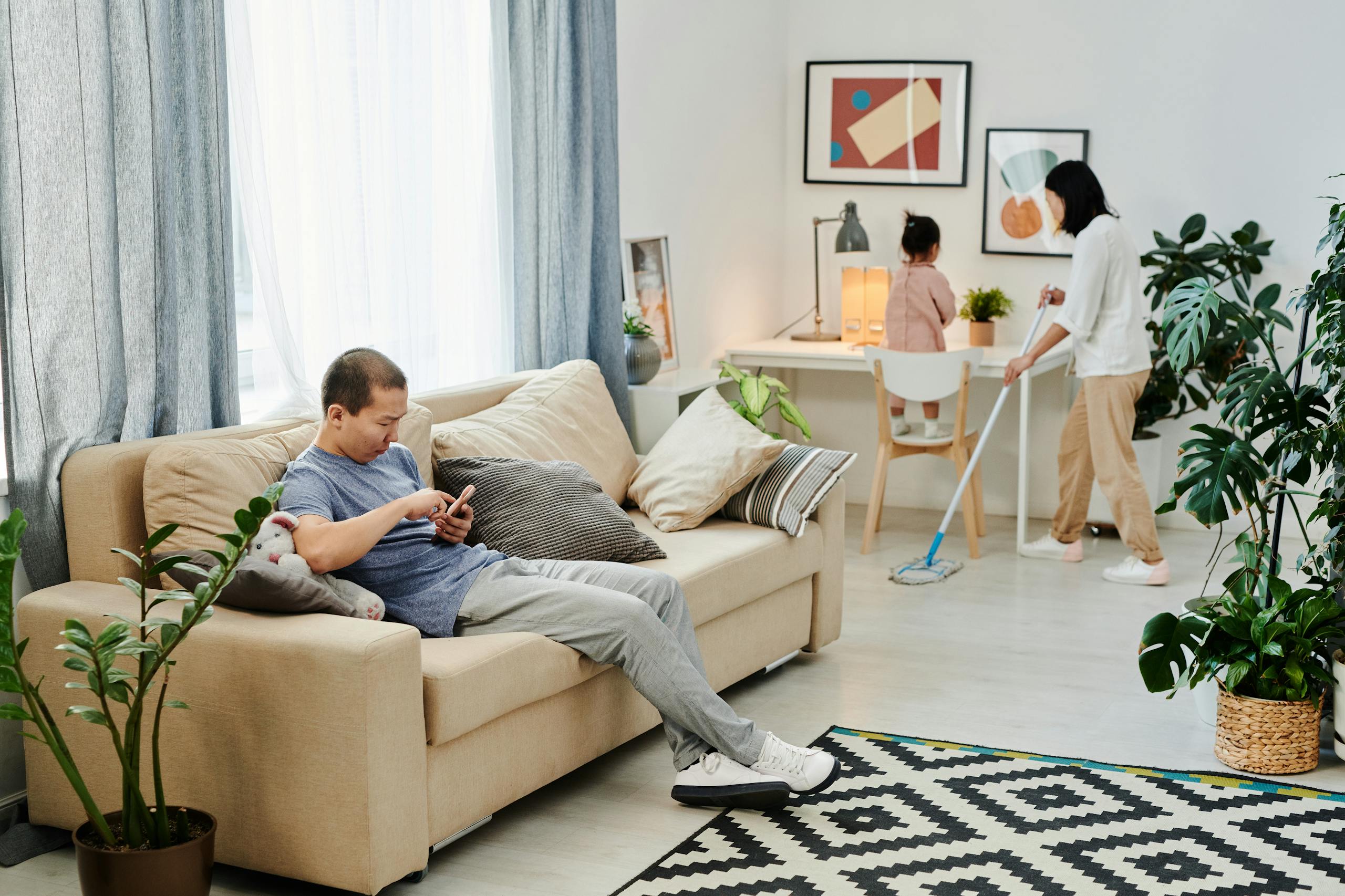 A family engages in daily chores, with a man relaxing and a woman mopping the floor.