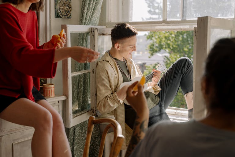 A group of young friends enjoying music and leisure time indoors with a ukulele by the window.