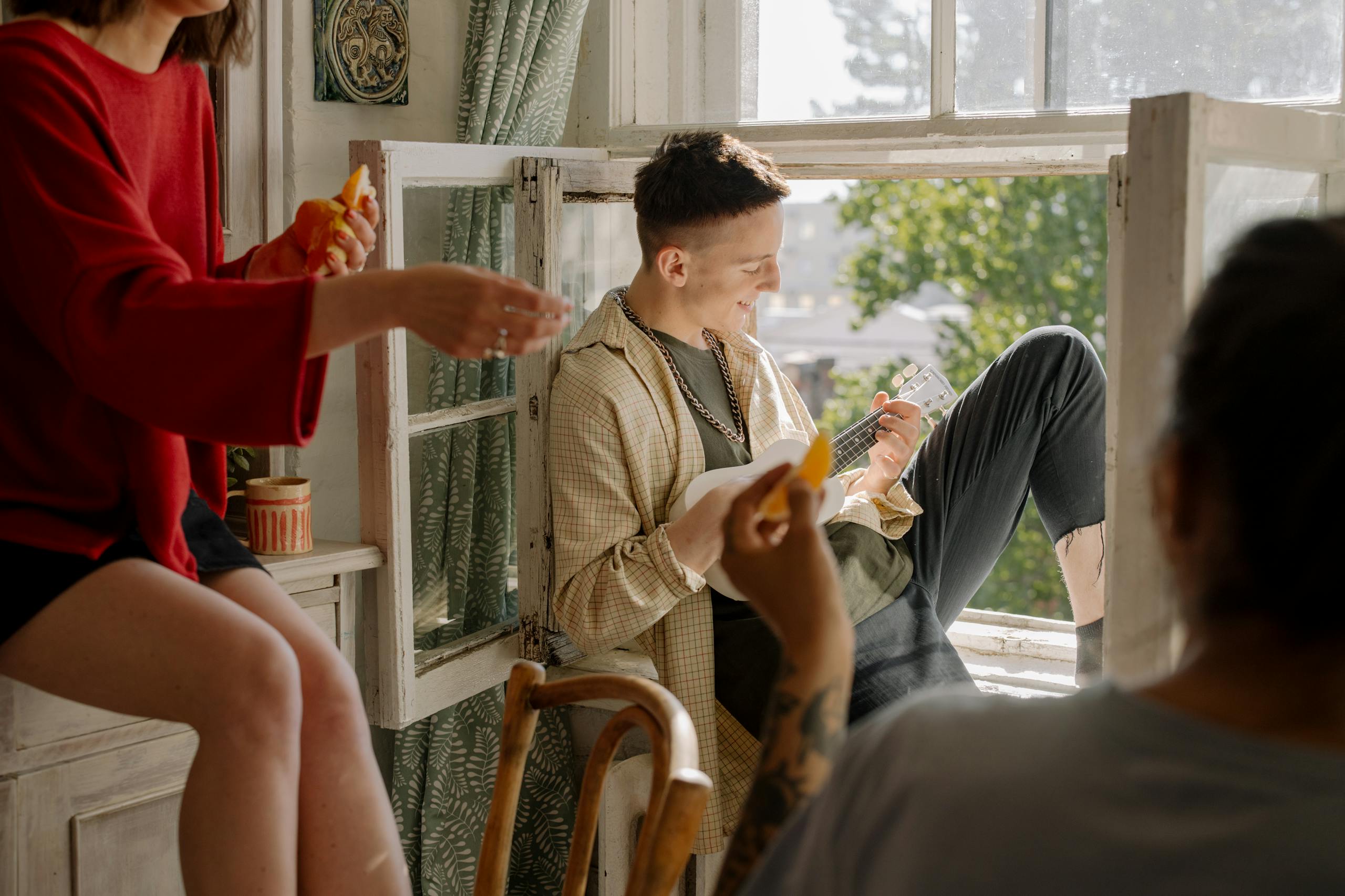 A group of young friends enjoying music and leisure time indoors with a ukulele by the window.