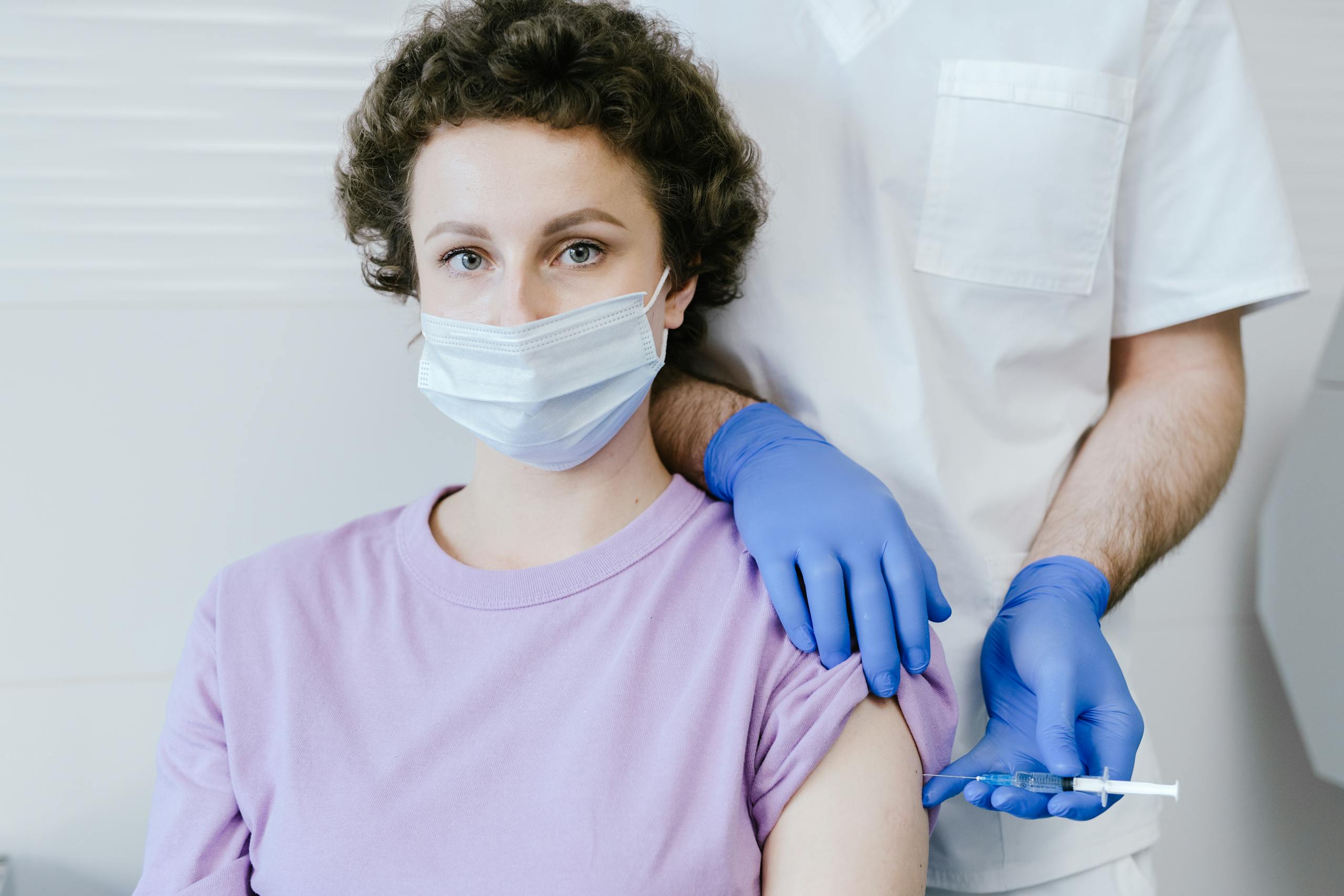A healthcare worker gives a vaccine injection to a masked patient, wearing gloves.