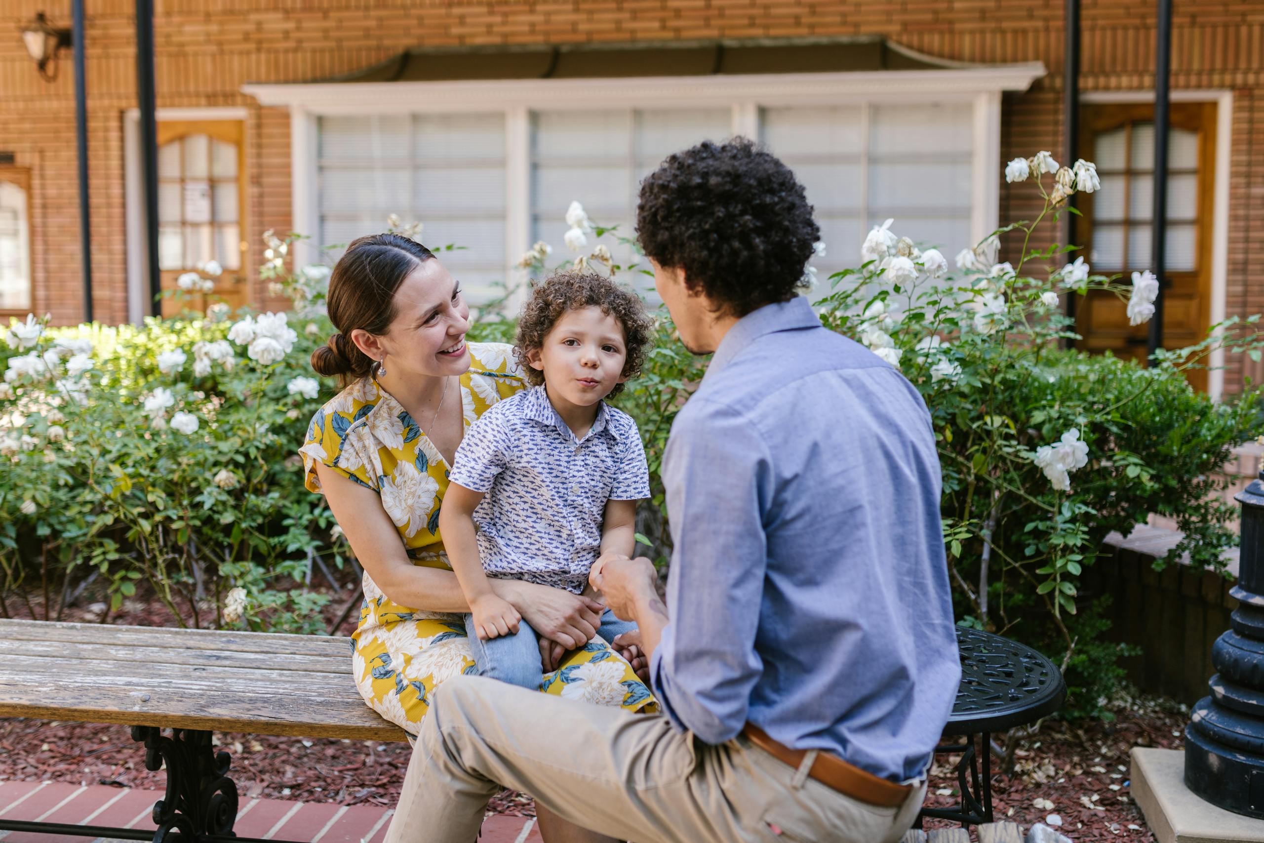 A joyful family moment outdoors with parents and child sitting on a bench surrounded by flowers.
