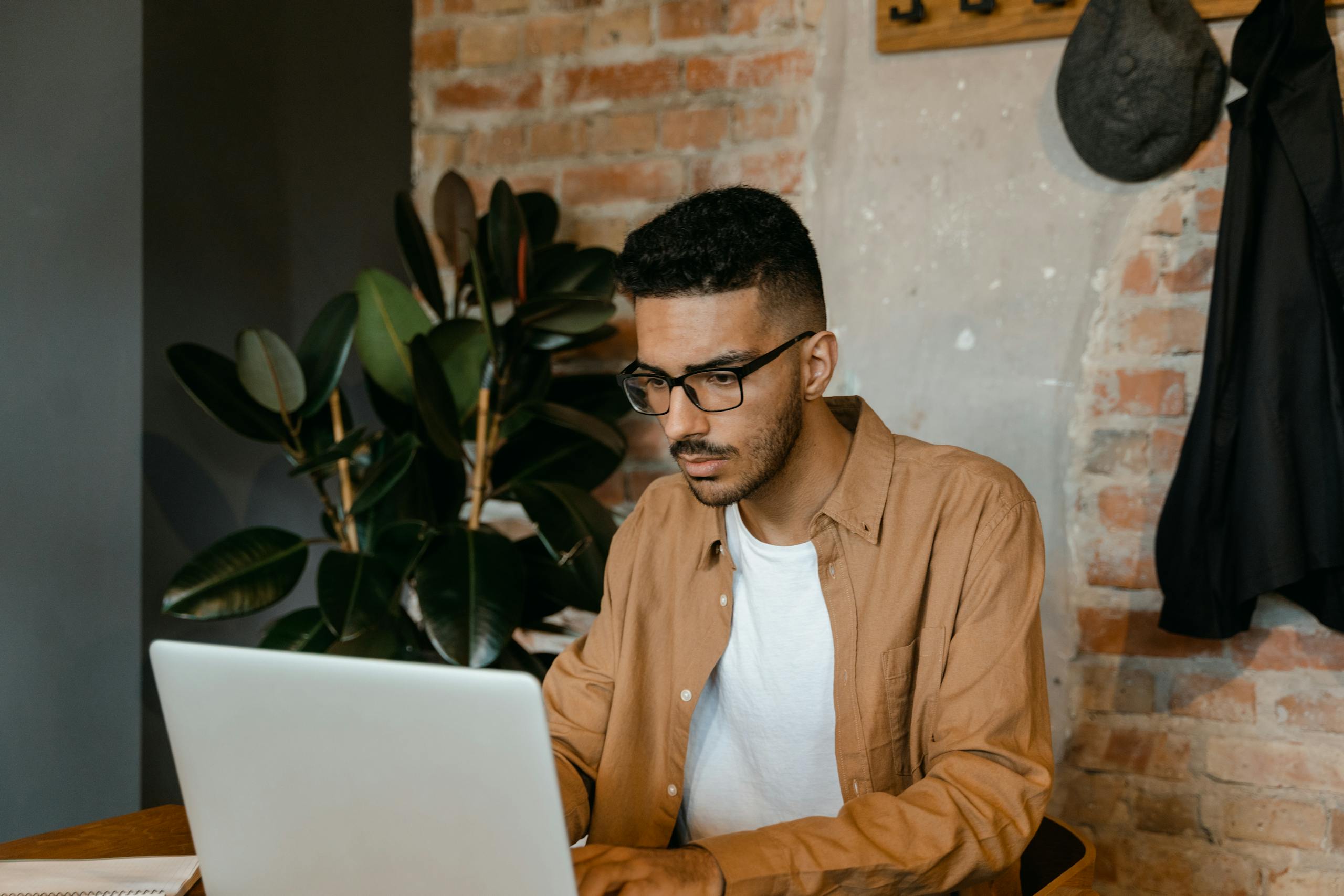 A man in glasses working on a laptop in a cozy cafe with brick walls and plants.