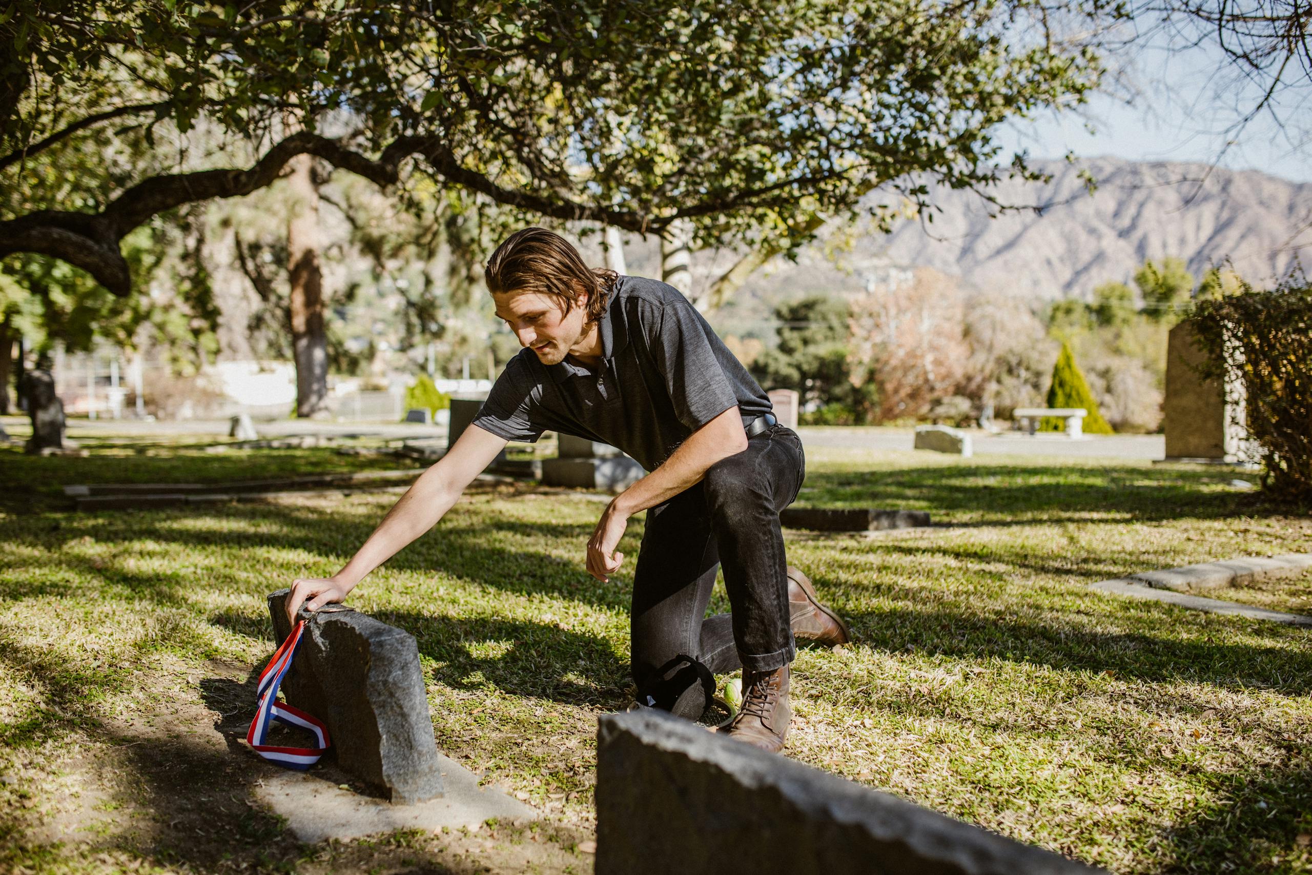 A man kneels beside a tombstone in a peaceful cemetery setting, symbolizing grief and remembrance.