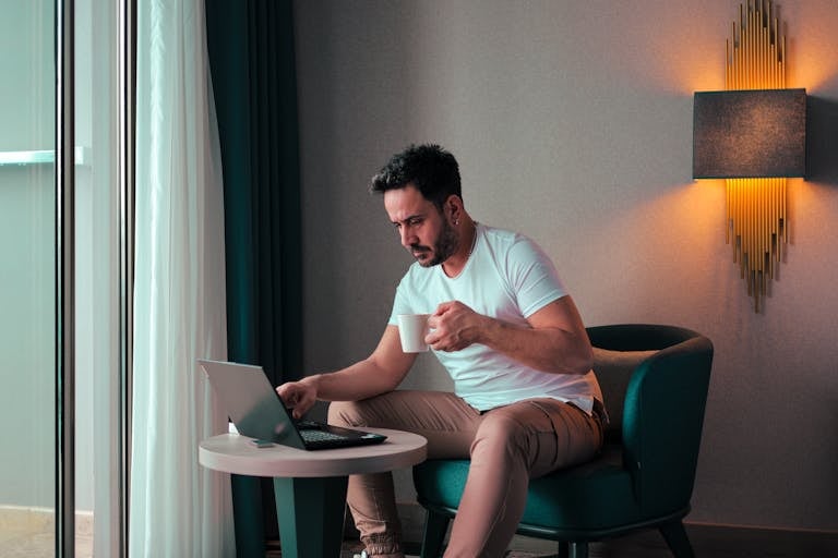 A man sits in a cozy hotel room in Cyprus, working on a laptop while enjoying a coffee.