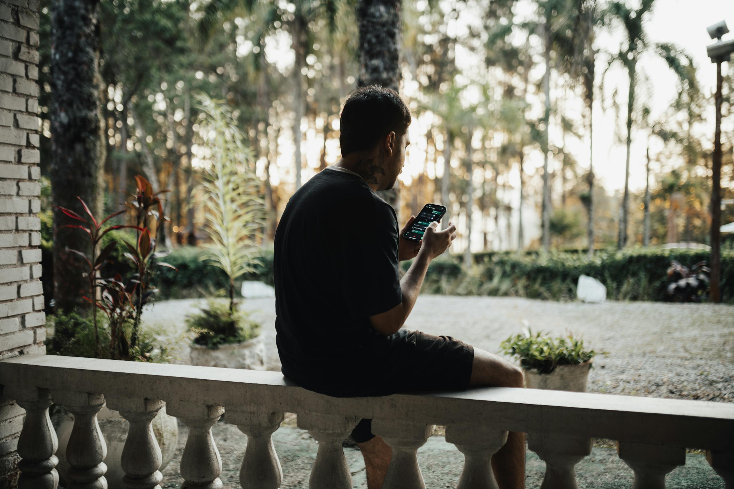 A man sitting on a patio bench using his smartphone, surrounded by lush greenery and palm trees.