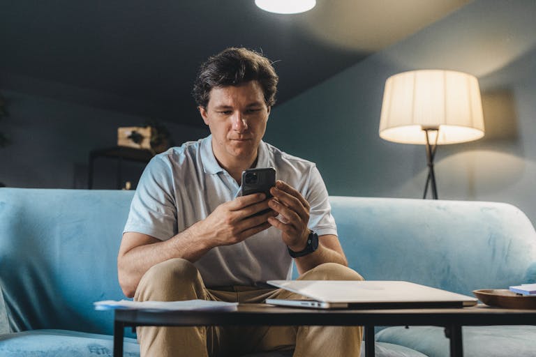 A man sitting on a sofa indoors using a smartphone under warm lighting.