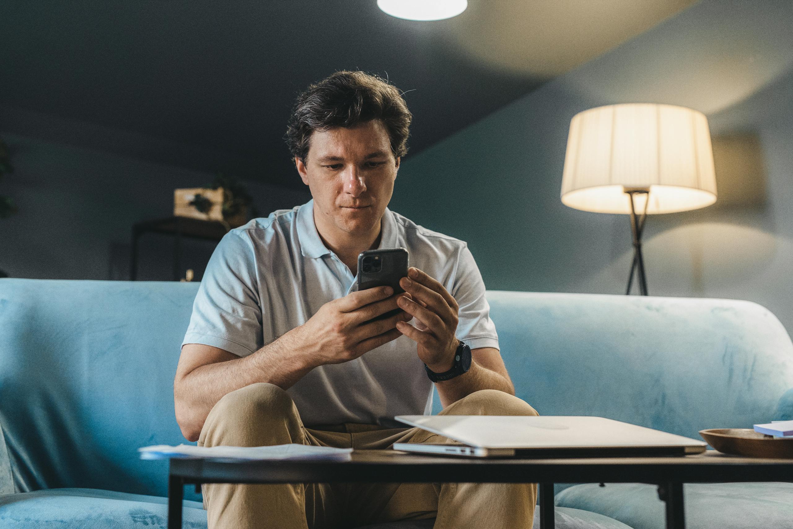 A man sitting on a sofa indoors using a smartphone under warm lighting.