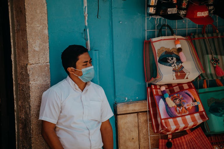 A man wearing a face mask leans against a wall in a vibrant Guanajuato market street scene.