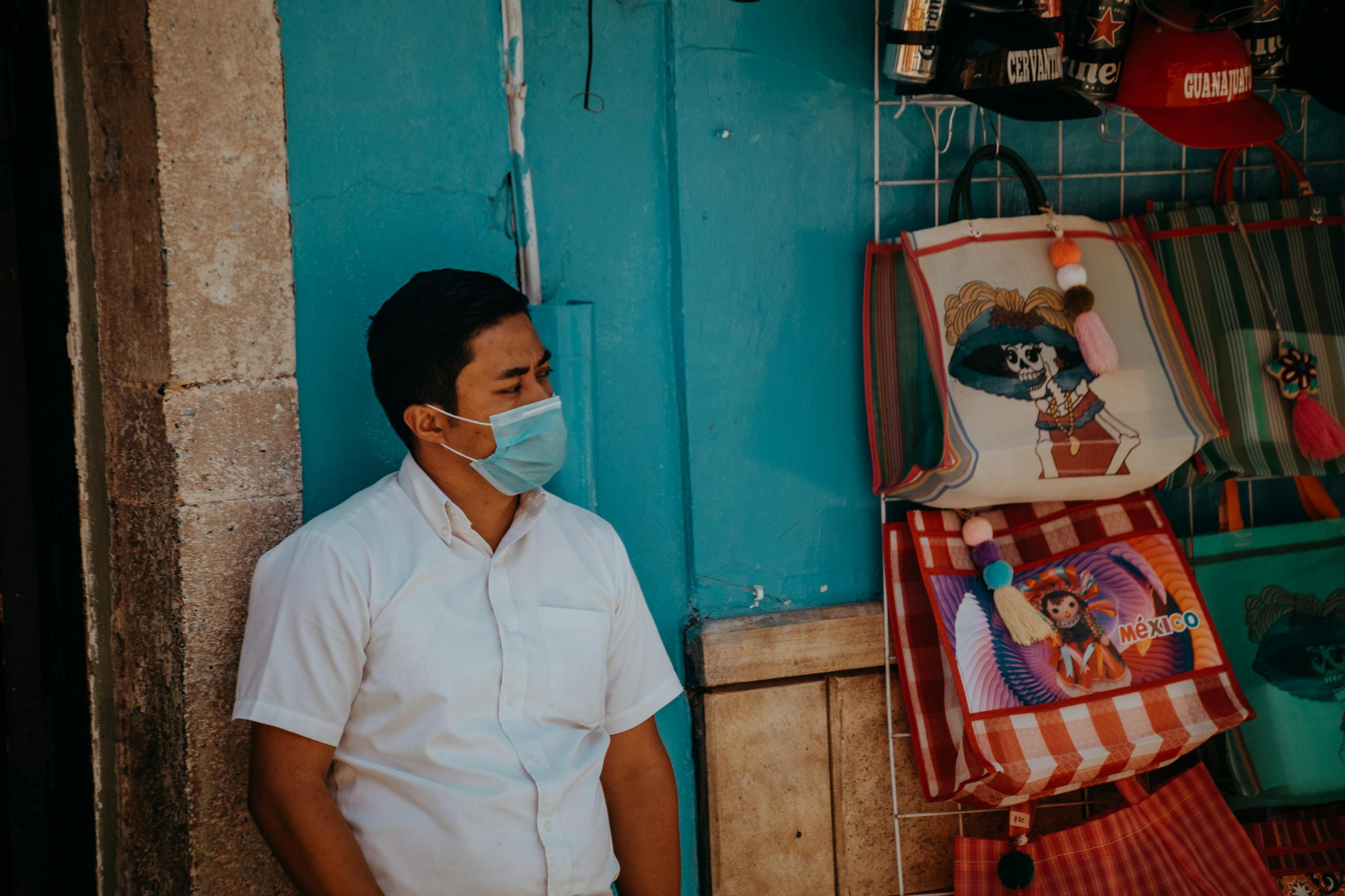 A man wearing a face mask leans against a wall in a vibrant Guanajuato market street scene.